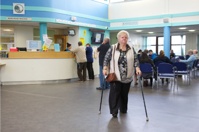 Older woman walking with sticks in the busy reception area of a GP surgery in the UK.